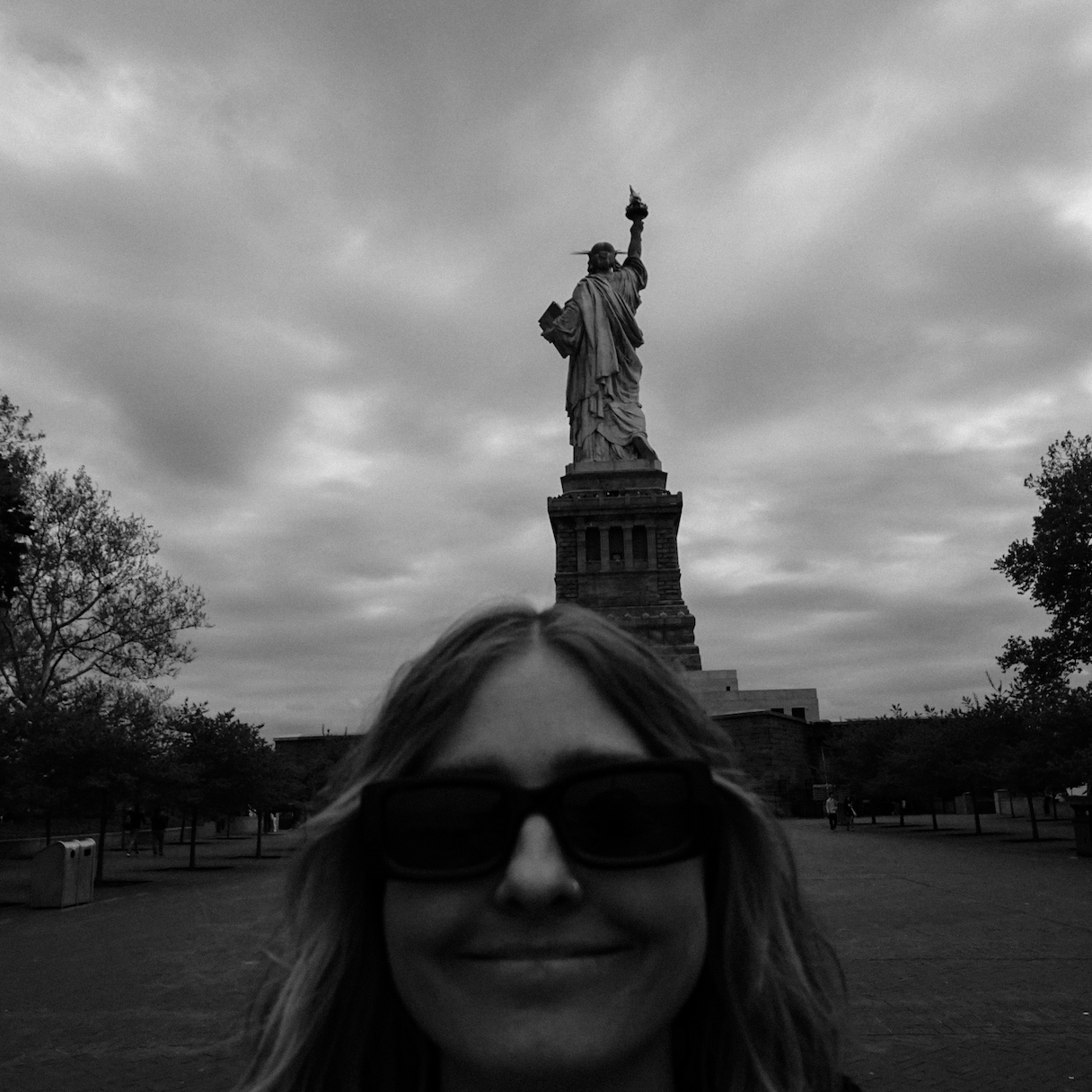 Alexa smiling in a plaza on Liberty Island behind the Statue of Liberty